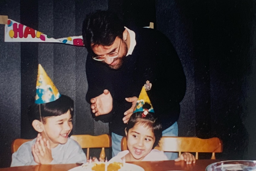 A Pakistani man celebrating a birthday with his two children. He's leaning forward, clapping, Kids wearing birthday hats