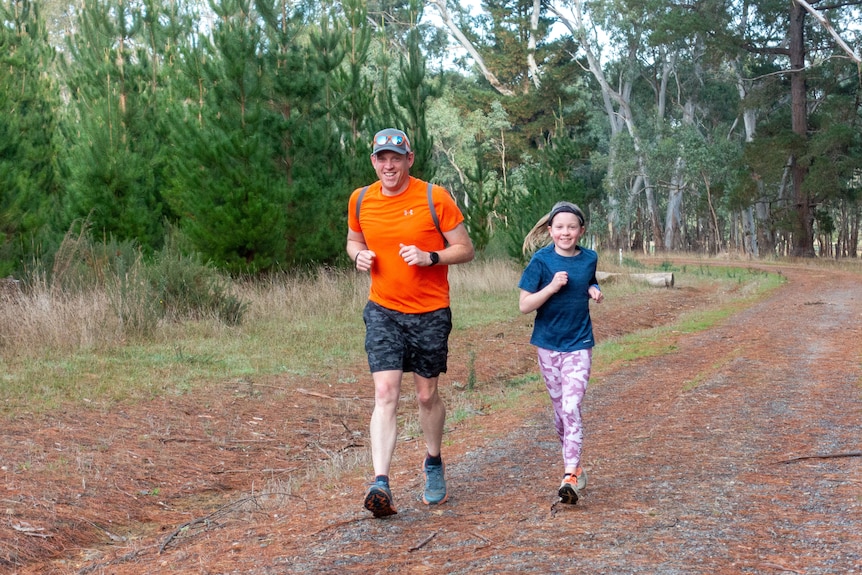 A man and young girl run along a dirt track surrounded by trees.