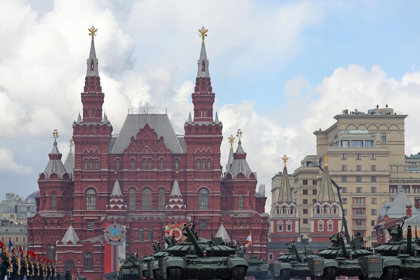 A line of tanks and soldiers on a street in frotn of tall buildings.