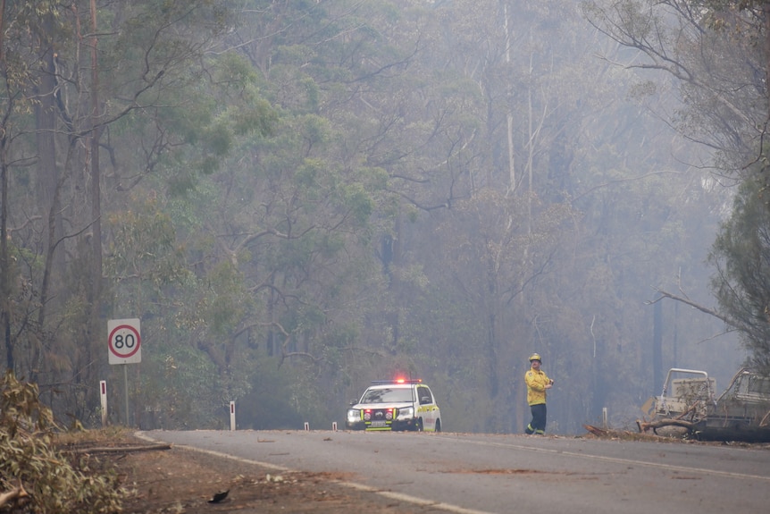 An emergency services worked in high vis clothing stands on a road near a car with flashing lights.