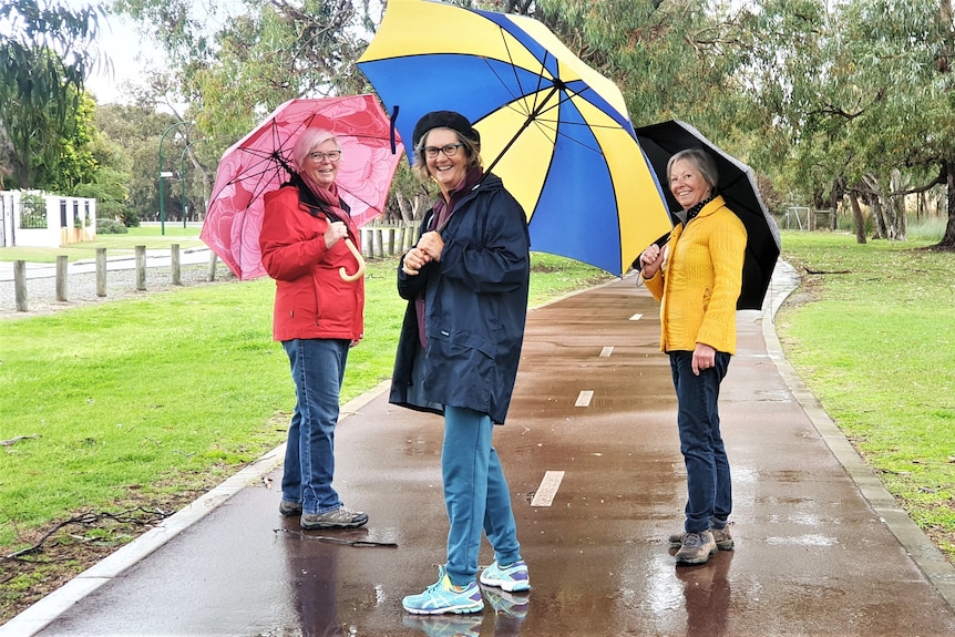 Three people hold colourful umbrellas in a park on a footpath.