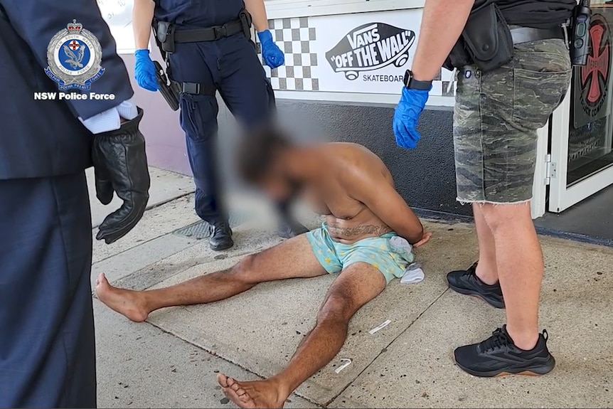 A shirtless man in shorts on the floor surrounded by police. His face is blurred out.