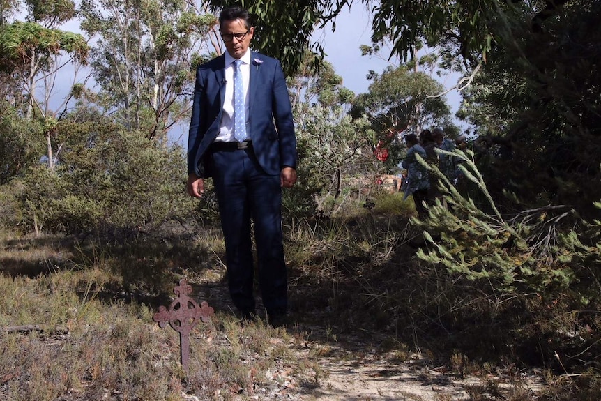 A man in a suit looks at a small iron cross in a bush setting.