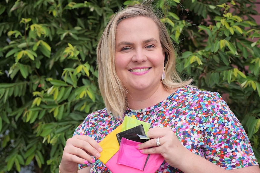 A lady with blond hair and a colourful shirt smiling and holding up period products.