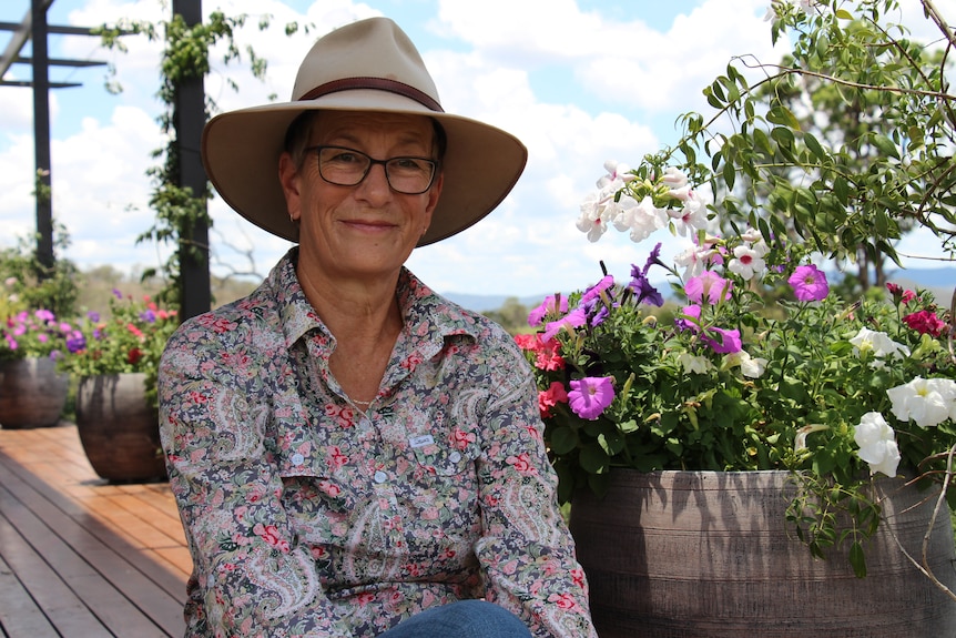 A woman in a button-up shirt and hat smiles while sitting on a deck, flowers behind her