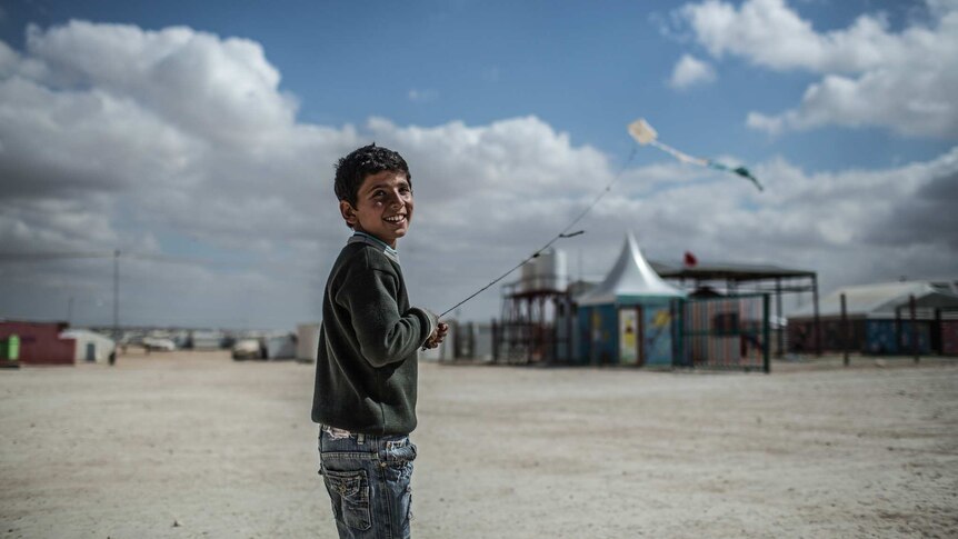 A boy, foreground, turning and smiling, with his kite flying in the distance in a refugee camp.
