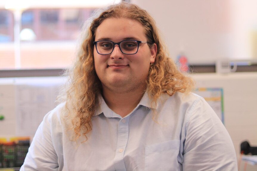 Dylan Botica, a young man with glasses and wearing a white shirt smiles, he has long curly blond hair.