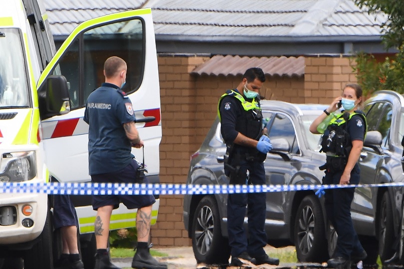 An ambulance is parked in a suburban street which has been blocked by police tape, as masked officers speak to a woman.