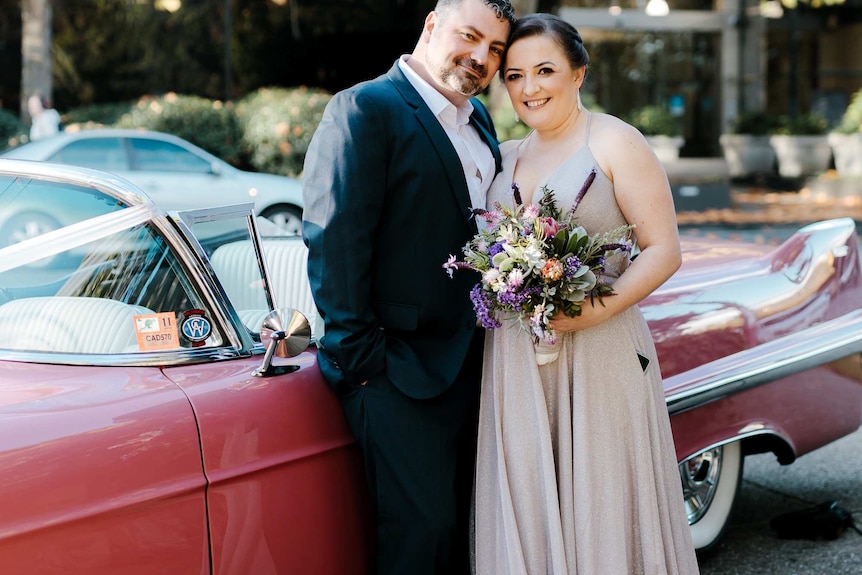 A woman in a long pale grey dress holds a bunch of flowers and stands close to a man in a suit with a red vintage car behind