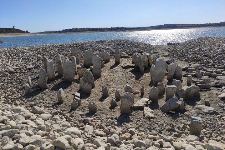 Dozens of large rocks arranged upright in a circle formation close to a river, surrounded by smaller stones.