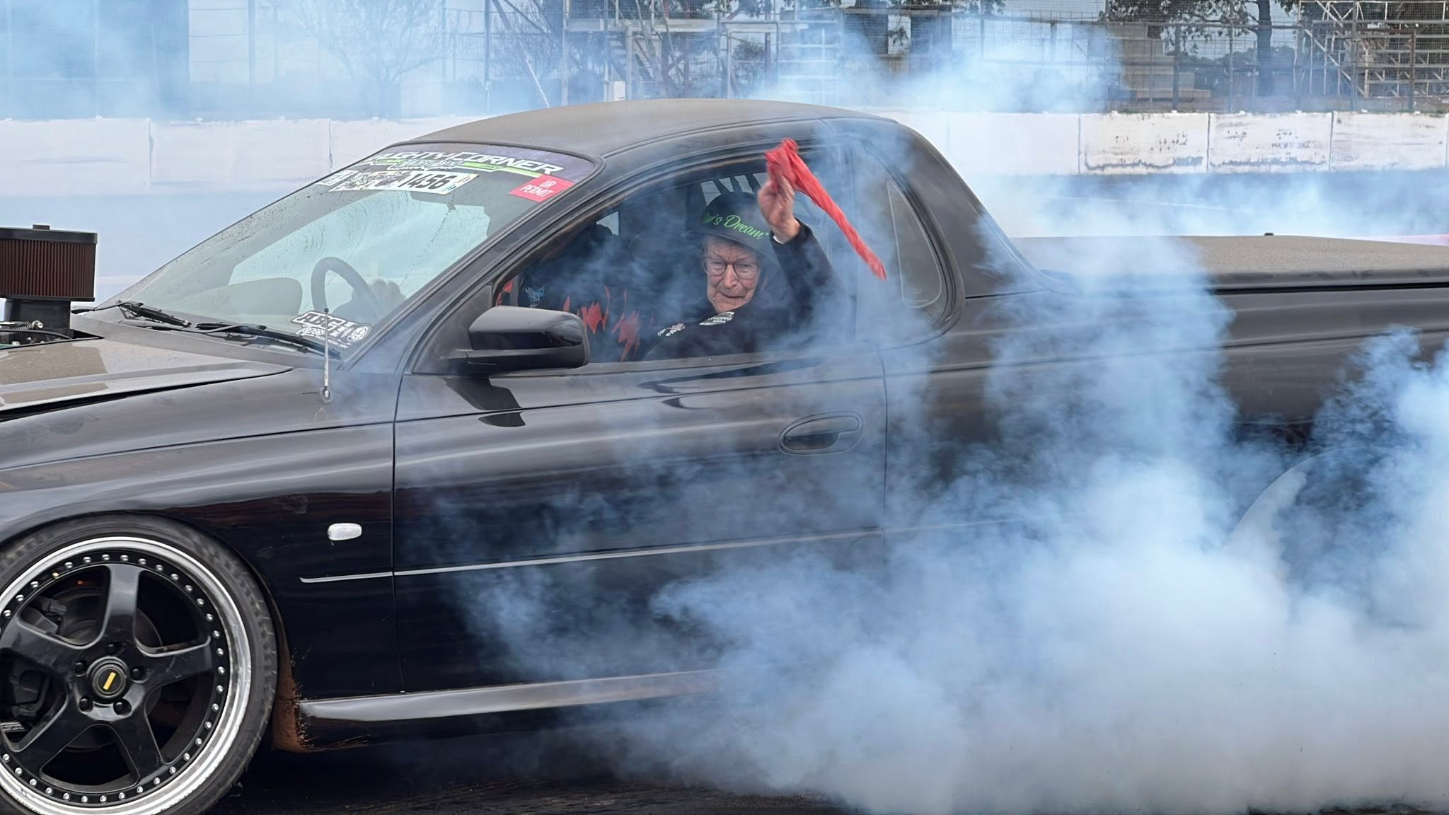 A woman with a helmet on waves an orange flag out a black ute with grey smoke coming out the back. 