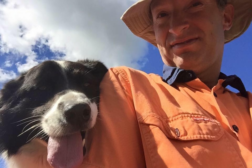 Mathew Dunbar, in a bright orange work shirt and hat sits to right of frame with a black and white working dog to his left