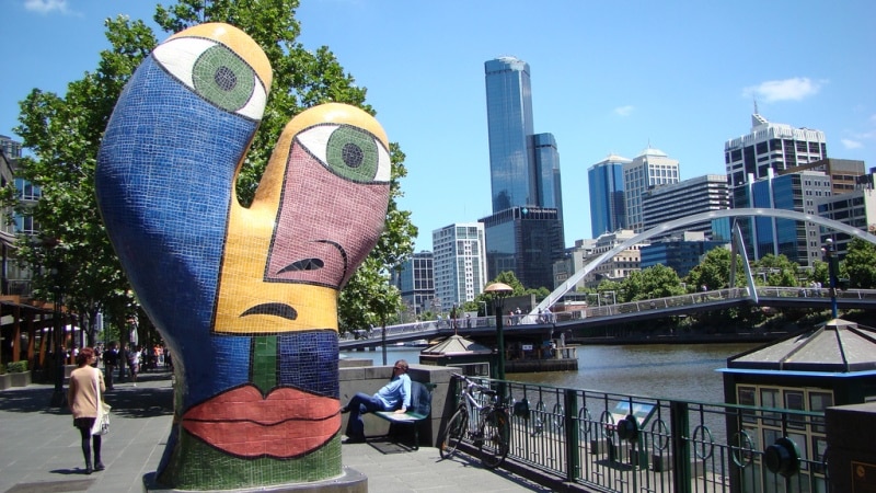 A large colourful mosaic statue of a face beside a city river.