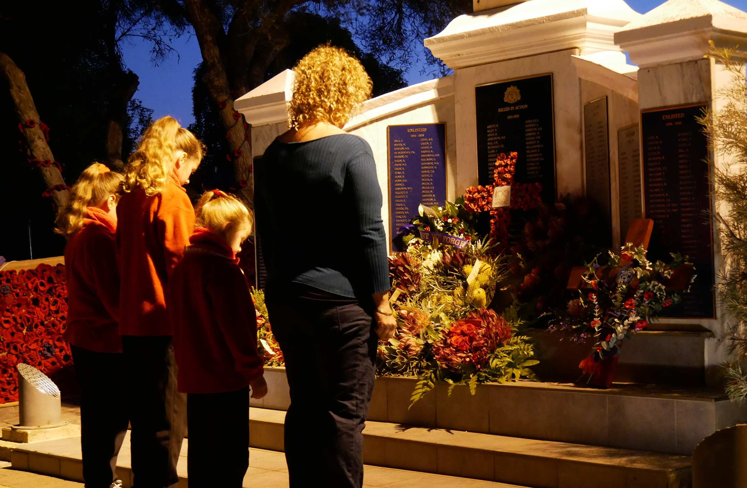 A woman and three girls in front on a war memorial and floral tributes in the pre-dawn light.