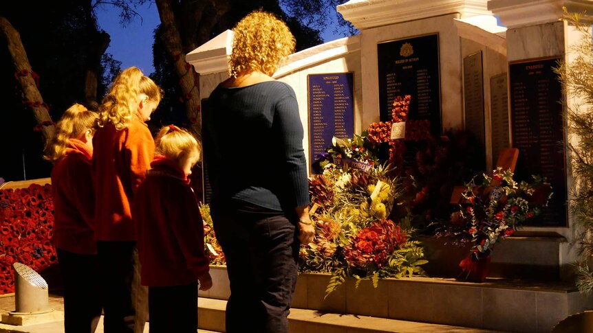 A woman and three girls in front on a war memorial and floral tributes in the pre-dawn light.