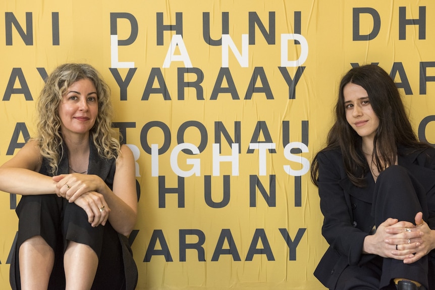 A woman with curly blonde hair and a woman with dark straight hair sit either side of a yellow and black text-based artwork.