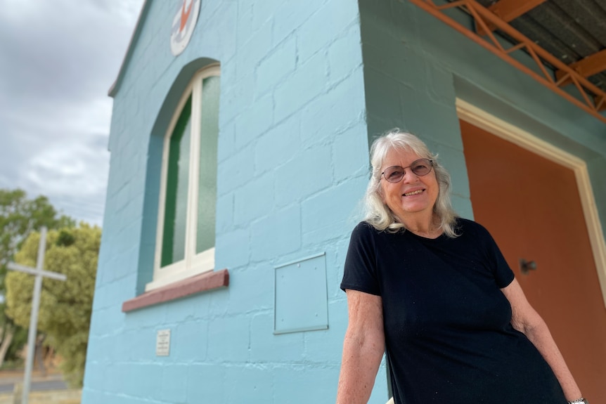 a woman with white hair and glasses smiling as she stands in front of a blue church