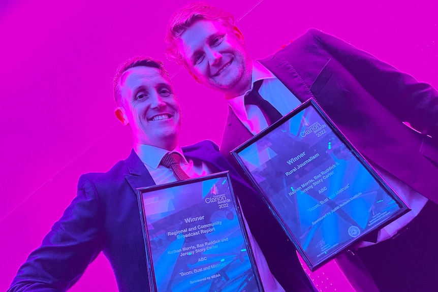 Two smiling men hold awards against a bright pink background