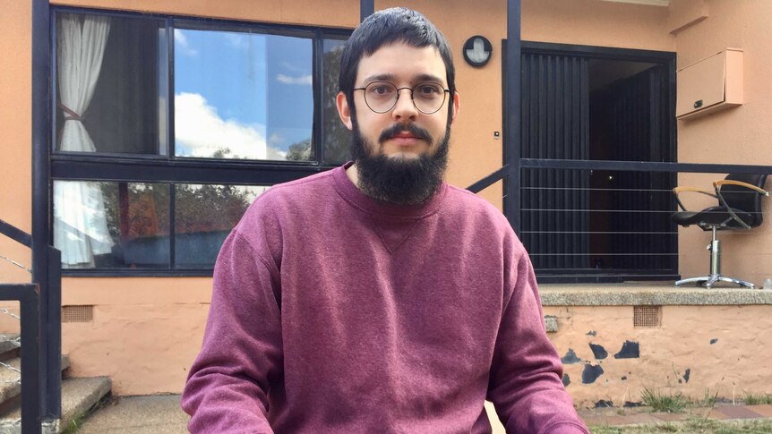 A young man with glasses and a beard sits outside his home.