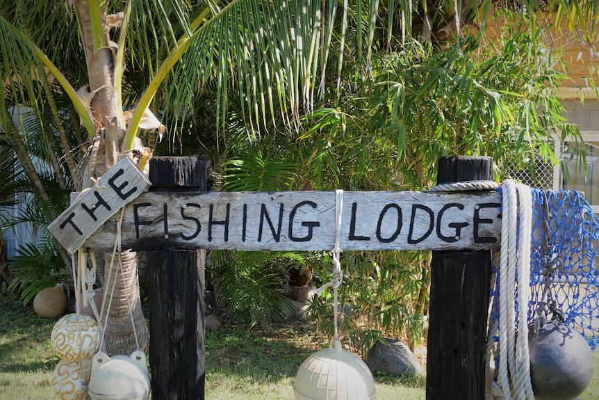 A fishing lodge wooden sign with netting and greenery in background.