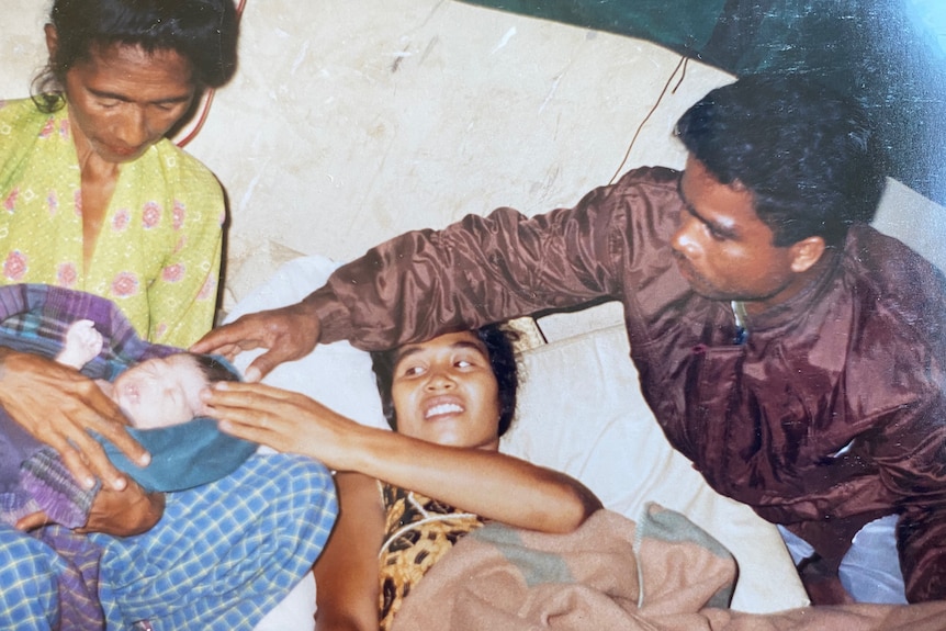 An older woman on the left holds a baby wrapped in blankets next to his mother and father who are lying next to them