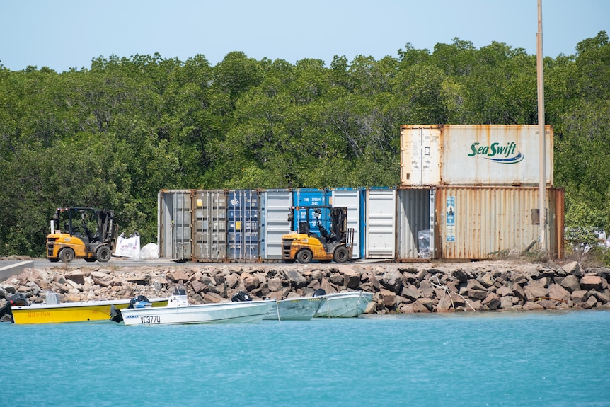 Shipping containers stacked alongside a wharf, with two small boats in the light blue water.