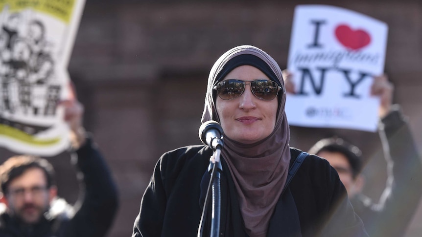 Linda Sarsour speaks at a rally, with protesters holding signs behind her.