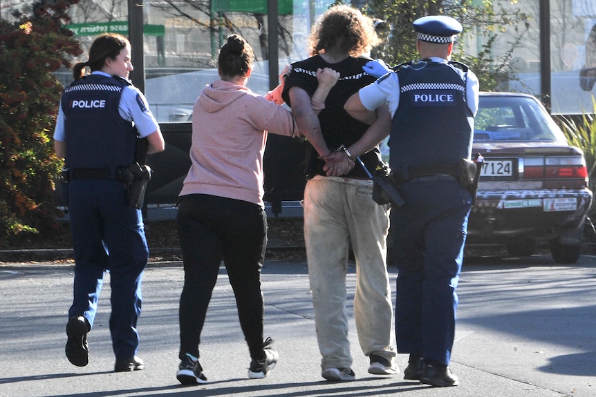 Two police officers anda  woman in casual clothing lead away an man in handcuffs, all of their backs to the camera. 