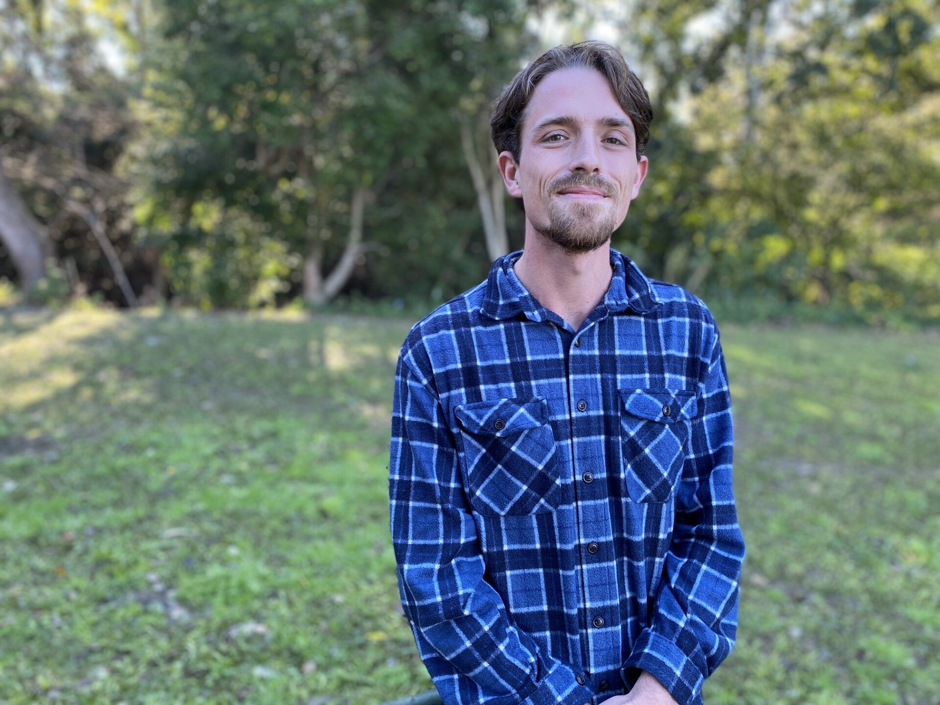 Man in checkered blue shirt with combed hair and small beard looking and smiling at camera 