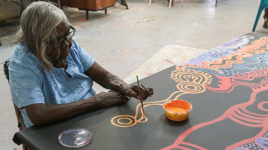 An elderly Indigenous woman paints an intricate design on a canvas.