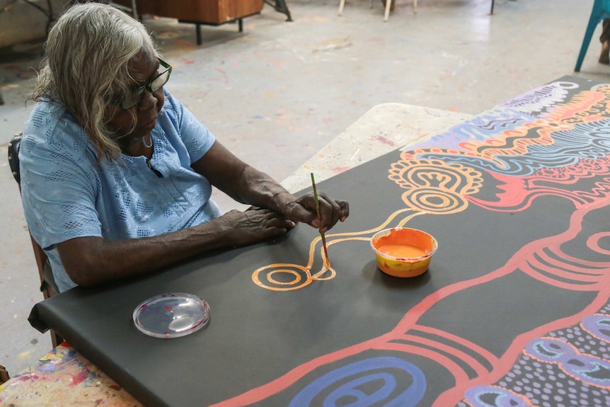 An elderly Indigenous woman paints an intricate design on a canvas.