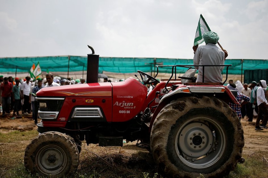 A man in a green turban holds a green flag as he sits on a red tractor with a group of people behind him in rural setting