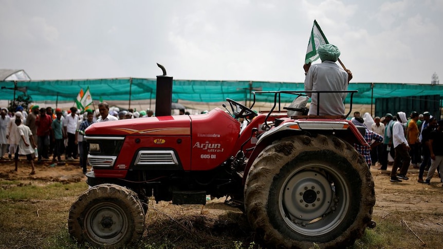 A man in a green turban holds a green flag as he sits on a red tractor with a group of people behind him in rural setting