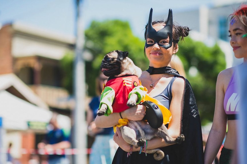 A woman dressed as Batman holds a dog dressed as Robin.