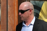 A bald man in a suit and sunglasses walks in the sunshine outside a building in Perth's CBD.