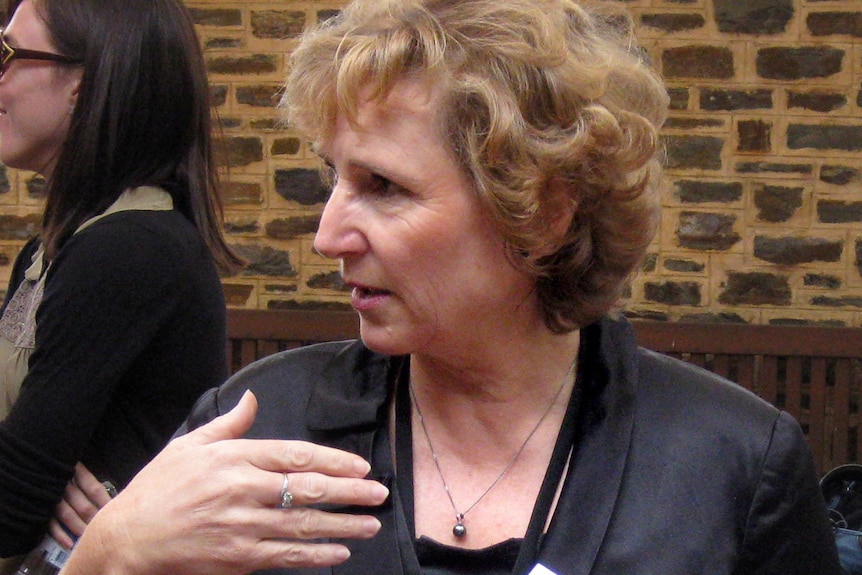 A woman with curly, short auburn hair looks to her right as she gesticulates with her right hand. She is wearing a black outfit.