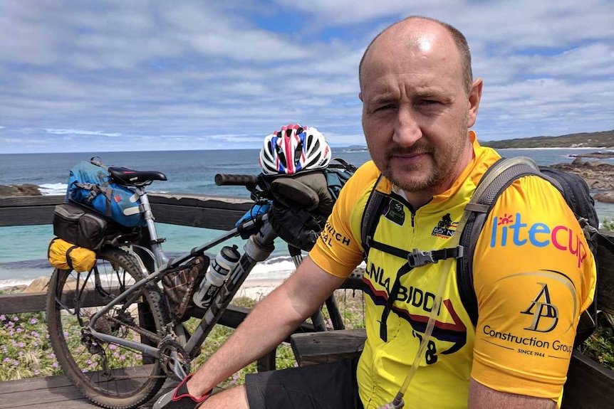 A man with a beard sitting on bench near the ocean, next to a bike.