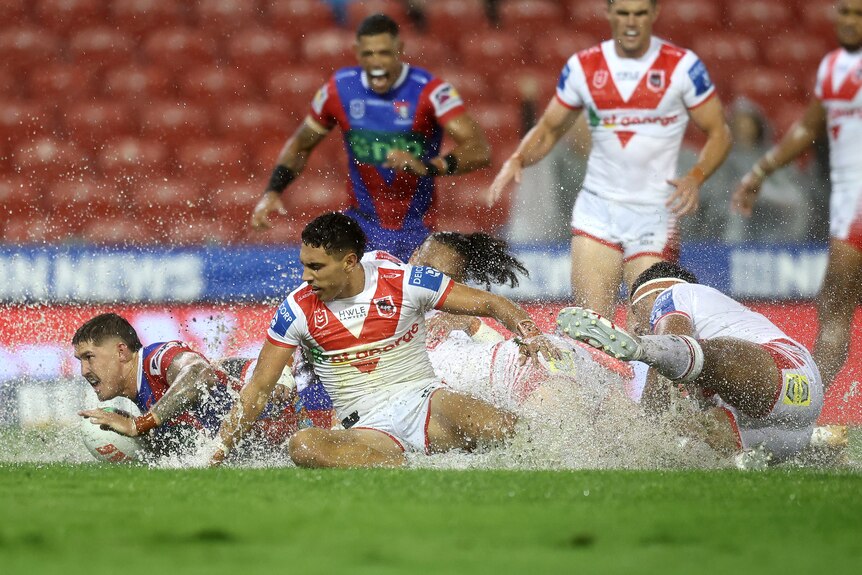 A Newcastle NRL player slides through a huge spray of water after scoring a try, with two Dragons defenders behind him.