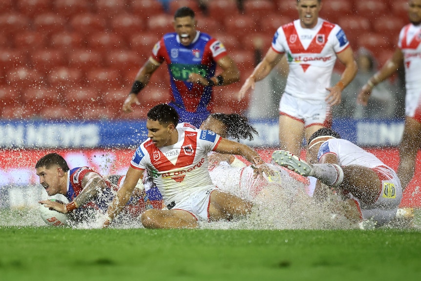 A Newcastle NRL player slides through a huge spray of water after scoring a try, with two Dragons defenders behind him.