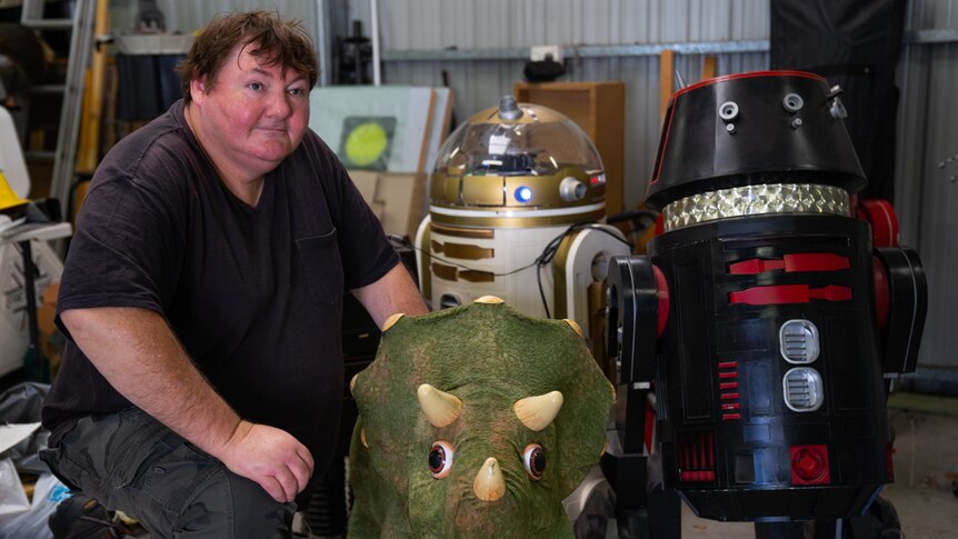 Robot artist Paul Aitken kneels alongside his robotic dinosaur and R2D2 droids.