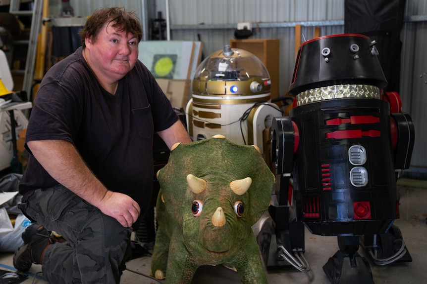 Robot artist Paul Aitken kneels alongside his robotic dinosaur and R2D2 droids.