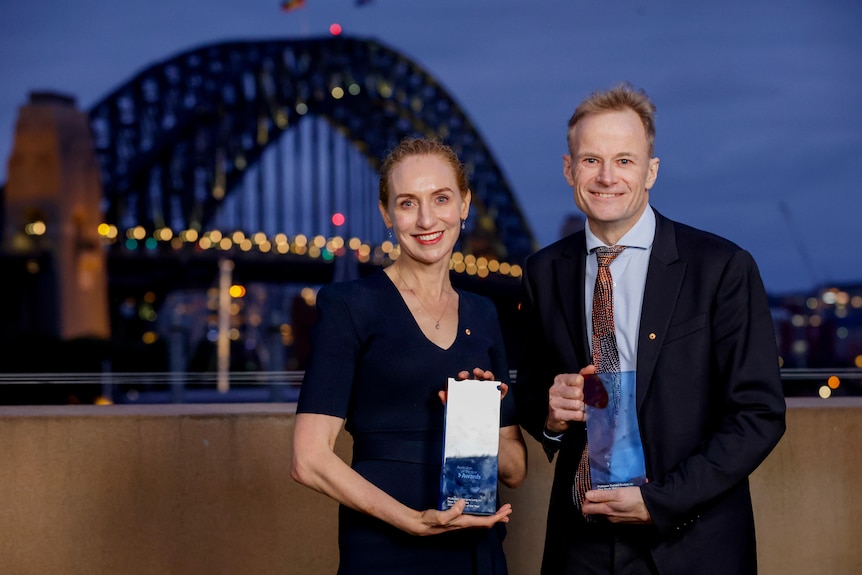 A man and a woman dressed smartly hold up glass awards with the Sydney Harbor Bridge in the background.