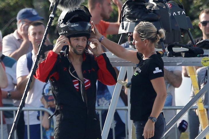 Franky Zapata puts his helmet on and as a blonde woman helps him. He wears a black and red suit. People watch.