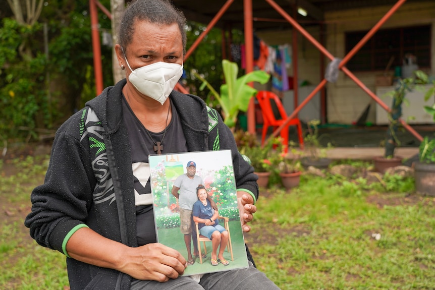 A woman in a face mask holds a photo of a man and a woman