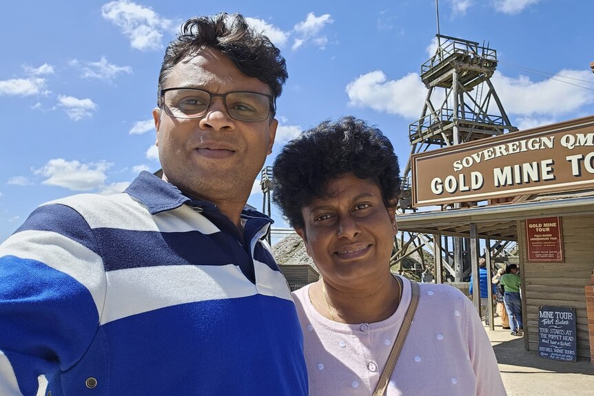 A smiling man and woman stand in front of a historic tourist attraction and take a selfie.