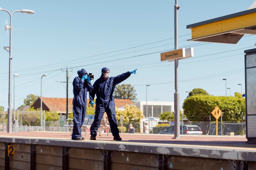 Two WA Police forensic officers in protective clothing stand on the platform at Queens Park train station.