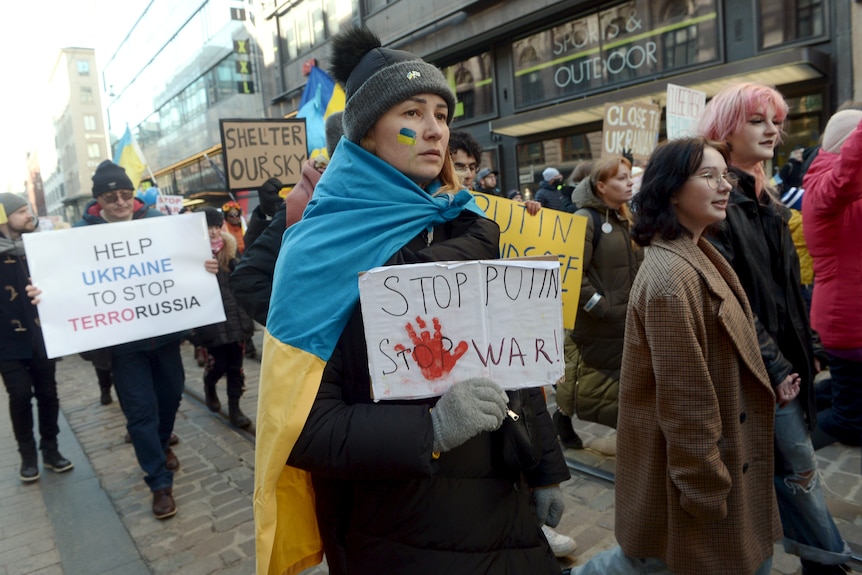 A woman wearing a ukrainian flag and beanie holds a sign that says stop Putin's war.