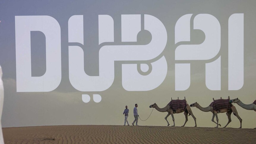 letters 'DUBAI' across poster of sanddunes and man leading camels. Man in traditional Arab dress in front of poster
