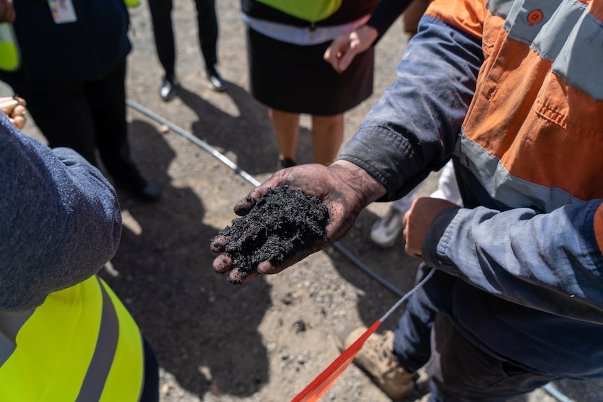A person in high vis holds a handful of a black charred looking substance, similar to soil.