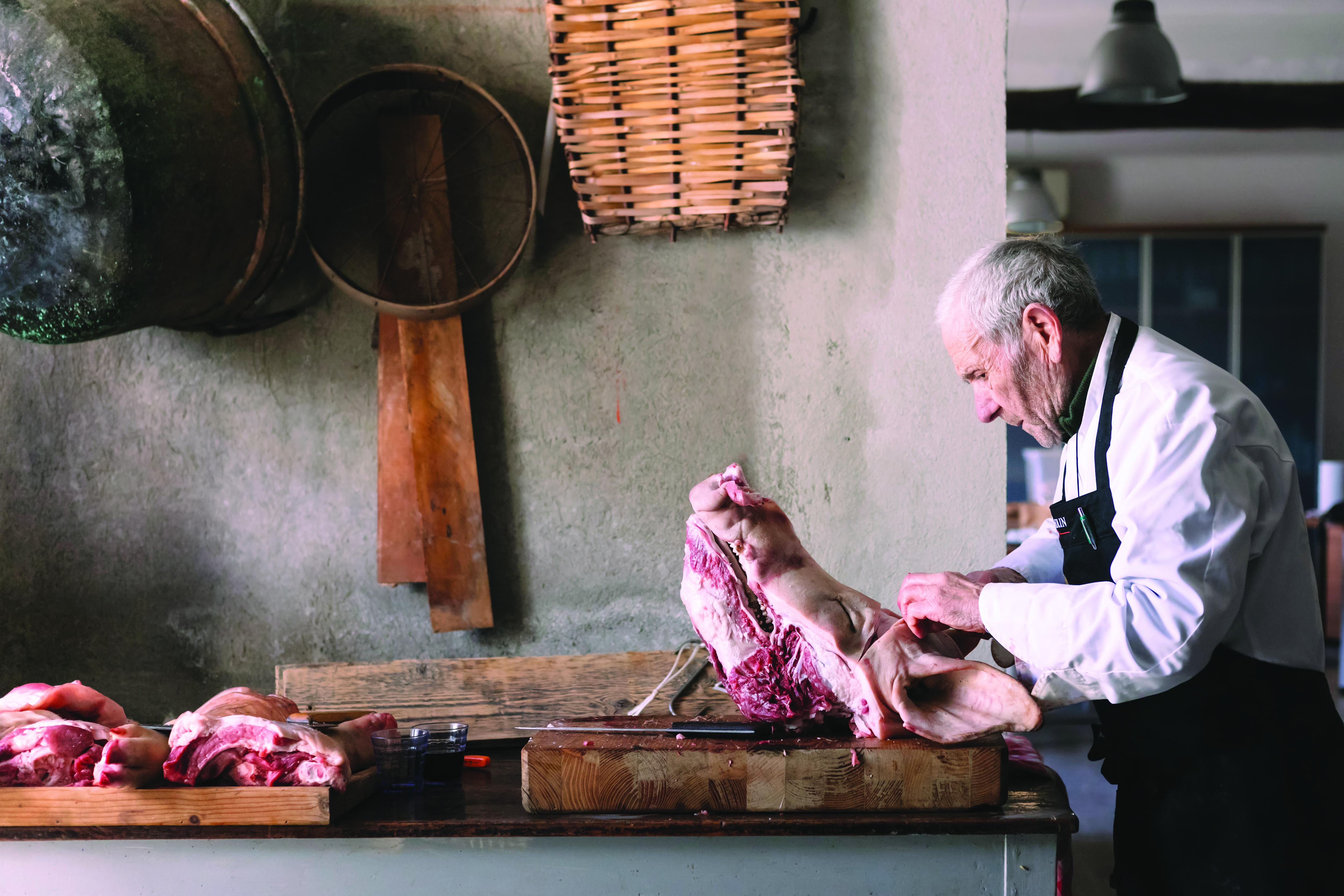 A man butchers a pig&#x27;s head in the middle of a rustic kitchen. 